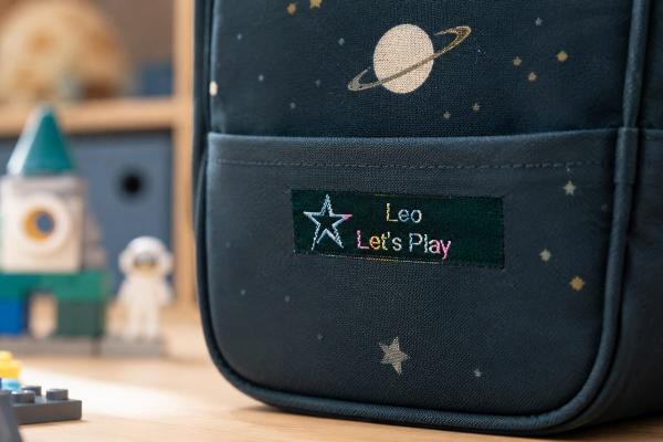 Moosgrünes Baumwoll-Etikett mit Regenbogen-Schrift auf Kinderrucksack