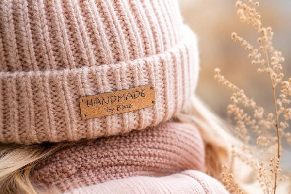 Rechteckiges hellbraunes Kunstlederetikett "Handmade by Bine" an rosa Strickmütze.